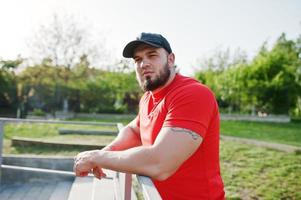 jeune homme musclé barbu brutal portant une chemise rouge, un short et une casquette au stade. photo