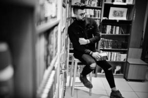 grand étudiant arabe intelligent, portant une veste en jean noir et des lunettes, à la bibliothèque avec un livre à portée de main. photo