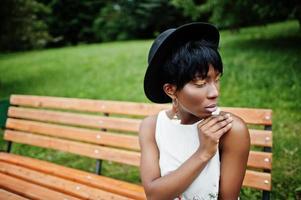 incroyable femme modèle afro-américaine en pantalon vert et chapeau noir posé sur un banc au parc. photo