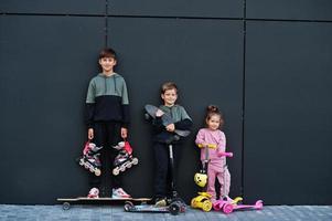 trois enfants en plein air contre un mur moderne noir. la famille sportive passe du temps libre à l'extérieur avec des scooters et des patins. photo