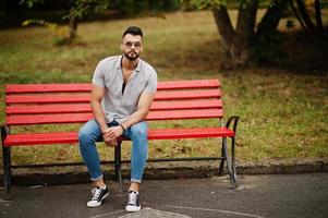 grand homme à la barbe arabe à la mode portant une chemise, un jean et des lunettes de soleil assis sur un banc rouge au parc. photo