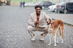 homme afro élégant en costume beige old school avec chien barzoï russe. jeune homme africain à la mode en veste décontractée sur torse nu. photo