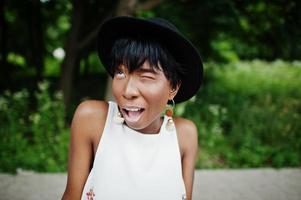 incroyable femme modèle afro-américaine en pantalon vert et chapeau noir posé au parc, clin d'oeil. photo