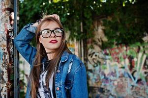 élégante fille hipster décontractée en jeans et lunettes contre un grand mur de graffitis. photo