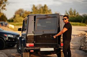 l'homme asiatique porte sur tout le noir posé près de la voiture suv, porte arrière ouverte. photo