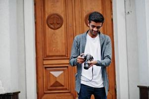 Un jeune photographe indien élégant porte une tenue décontractée posée en plein air avec un appareil photo reflex numérique à portée de main.