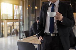 homme d'affaires tenant une tasse de café et regardant ailleurs tout en se tenant à l'extérieur avec un café en arrière-plan, redémarrant après une dure journée de travail. photo