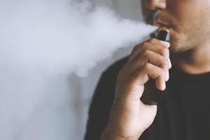 photo portrait jeune homme avec barbe tenir et fumer sa cigarette électronique soufflant un flux de fumée se propager. noir foncé, fond gris