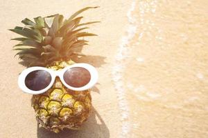 ananas avec des lunettes de soleil prendre un bain de soleil sur une plage tropicale avec une vague photo