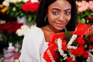 belle fille afro-américaine en robe blanche tendre avec bouquet de fleurs dans les mains debout sur fond floral dans le magasin de fleurs.fleuriste femme noire. photo
