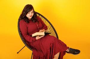 jolie femme sud-asiatique en robe rouge foncé posée au studio sur fond jaune assis sur une chaise avec un livre. photo