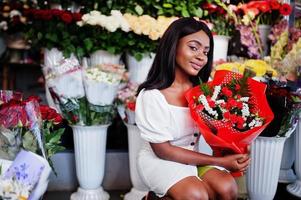 belle fille afro-américaine en robe blanche tendre avec bouquet de fleurs dans les mains debout sur fond floral dans le magasin de fleurs.fleuriste femme noire. photo