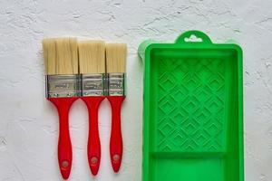 trois pinceaux avec poignées rouges et bac à peinture vert sur fond de béton blanc. outils et accessoires pour la rénovation domiciliaire. vue de dessus photo