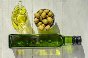 saucière en verre avec huile d'olive extra vierge, olives vertes fraîches et bouteille sur table en bois.