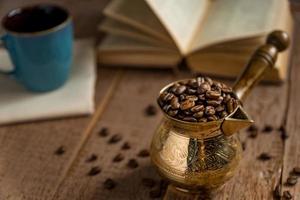 grains de café torréfiés frais dans une cafetière turque traditionnelle cezve livre ouvert et tasse sur une table en bois. photo