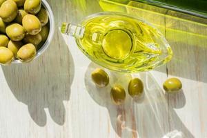 saucière en verre avec huile d'olive extra vierge, olives vertes fraîches et bouteille sur table en bois.