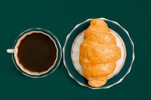 vue de dessus d'une tasse de café noir et d'un croissant sur une assiette sur fond vert foncé. petit-déjeuner du matin à la française. photo