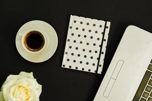 table de bureau à plat, vue de dessus. espace de travail avec ordinateur portable, rose blanche, journal à pois et tasse à café sur fond noir. photo