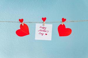 épingles de coeurs d'amour rouges accrochées à un cordon naturel sur fond bleu. inscription de bonne fête des mères sur une feuille de papier. photo