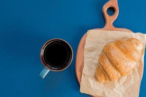 vue de dessus d'une tasse de café noir et d'un croissant sur une planche à découper et du papier kraft sur fond bleu avec espace de copie. petit-déjeuner du matin à la française.
