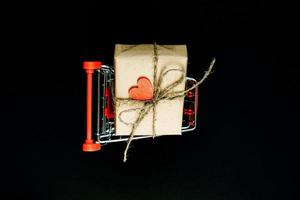 coffret cadeau artisanal enveloppé dans du papier kraft avec coeur en bois rouge et corde dans le panier de jouets, sur fond noir. vue de dessus, mise à plat photo