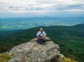 Gros homme asiatique assis sur une falaise rocheuse et méditation sur la montagne khao luang dans le parc national de ramkhamhaeng, province de sukhothai en thaïlande photo