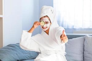 belle jeune femme avec un masque facial sur son visage tenant des tranches de concombre frais. soins et traitement de la peau, spa, beauté naturelle et concept de cosmétologie photo