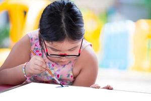 une fille asiatique fait de la peinture d'art sur du papier blanc. l'enfant porte des lunettes. les enfants mignons font de l'artisanat. enfant étudie à la maison. équipe de basket-ball de dessin animé de dessin féminin. dame âgée de 5-6 ans. favoriser l'imaginaire. photo