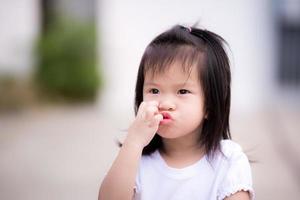 une fille heureuse utilise sa main pour se gratter le visage et faire une bouche plissée de bonne humeur. enfant de 3 ans porter une chemise blanche pour se promener les soirs d'été ou de printemps devant la maison. photo