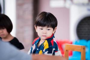 adorable garçon asiatique assis sur une chaise en bois. visage de bébé triste. enfant âgé de 2 ans, portant une chemise colorée. photo