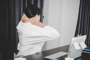 une jeune femme asiatique est assise au travail et a des douleurs de tension dans le cou. concept office syndrome mal de dos douleur due à une maladie professionnelle, femme ayant des douleurs musculaires à cause de l'utilisation d'un ordinateur portable, mal de dos. photo