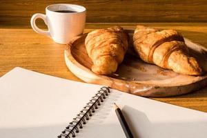 croissant avec tasse à café, cahier et crayon sur fond de table en bois. photo