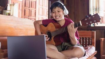 un mignon garçon asiatique portant des écouteurs des appels vidéo apprenant de la musique et écrivant des chansons en ligne sur son ordinateur portable à l'aide d'une guitare acoustique heureusement à la maison concept de créateurs d'enfants photo