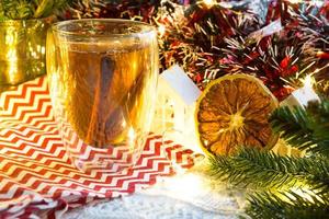 gobelet en verre transparent à double paroi avec thé chaud et bâtons de cannelle sur la table avec décor de noël et petite maison. ambiance nouvel an, tranche d'orange séchée, guirlande, branche d'épicéa, cosy
