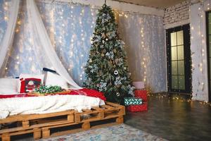 décor de noël dans la chambre dans le style loft. un lit à baldaquin fait de palettes en bois avec des draps blancs et des couvertures et des oreillers rouges. guirlandes d'épicéa et d'or, fenêtre illuminée. nouvel An. photo