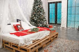 décor de noël dans la chambre dans le style loft. un lit à baldaquin fait de palettes en bois avec des draps blancs et des couvertures et des oreillers rouges. guirlandes d'épicéa et d'or, fenêtre illuminée. nouvel An. photo