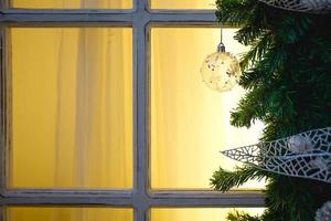 des décorations de noël faites de branches d'épinette et de boules et rubans transparents blancs encadrent la fenêtre avec cadre en bois et lumière jaune. ambiance festive du nouvel an, confort de la maison. espace pour le texte photo