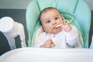 un bébé affamé ronge une cuillère en plastique à table sur une chaise haute. poussées dentaires, caprices, démangeaisons des gencives, introduction d'aliments complémentaires photo