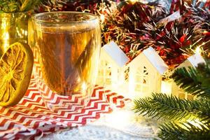 gobelet en verre transparent à double paroi avec thé chaud et bâtons de cannelle sur la table avec décor de noël et petite maison. ambiance nouvel an, tranche d'orange séchée, guirlande, branche d'épicéa, cosy photo