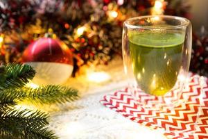 gobelet en verre transparent à double paroi avec thé matcha japonais sur table avec décor de noël. ambiance de nouvel an, guirlande et guirlande, branche d'épicéa, confortable, couverture tricotée, ballon, serviette rayée photo