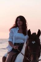 femme en vêtements d'été aime monter à cheval sur une belle plage de sable au coucher du soleil. mise au point sélective photo