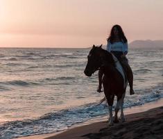 femme en vêtements d'été aime monter à cheval sur une belle plage de sable au coucher du soleil. mise au point sélective photo