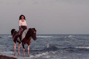 femme en vêtements d'été aime monter à cheval sur une belle plage de sable au coucher du soleil. mise au point sélective photo