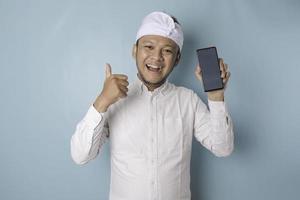 un homme balinais excité portant un bandeau udeng ou traditionnel et une chemise blanche donne le pouce en l'air geste d'approbation tout en tenant un smartphone, isolé sur fond bleu photo