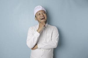 portrait d'un jeune homme balinais réfléchi portant un bandeau udeng ou traditionnel et une chemise blanche regardant de côté tandis que son doigt sur son menton isolé sur fond bleu photo