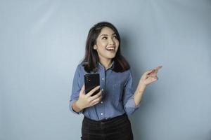 femme asiatique excitée portant une chemise bleue pointant vers l'espace de copie à côté d'elle tout en tenant un smartphone, isolée par un fond bleu photo