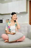 Une femme vietnamienne calme se repose sur un canapé et profite d'un son de bonne qualité dans les écouteurs du téléphone portable photo