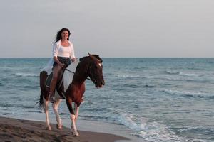 femme en vêtements d'été aime monter à cheval sur une belle plage de sable au coucher du soleil. mise au point sélective photo