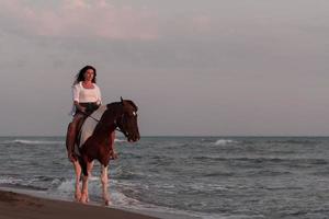 femme en vêtements d'été aime monter à cheval sur une belle plage de sable au coucher du soleil. mise au point sélective photo