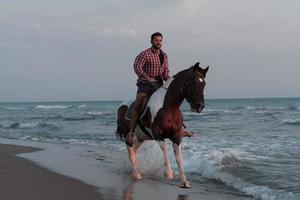 un homme moderne en vêtements d'été aime monter à cheval sur une belle plage de sable au coucher du soleil. mise au point sélective photo